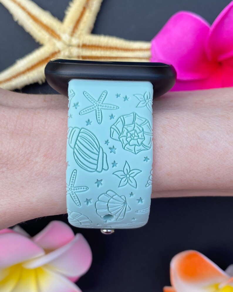 May include: A light blue silicone Apple Watch band with a seashell and starfish design. The band is on a person's wrist.
