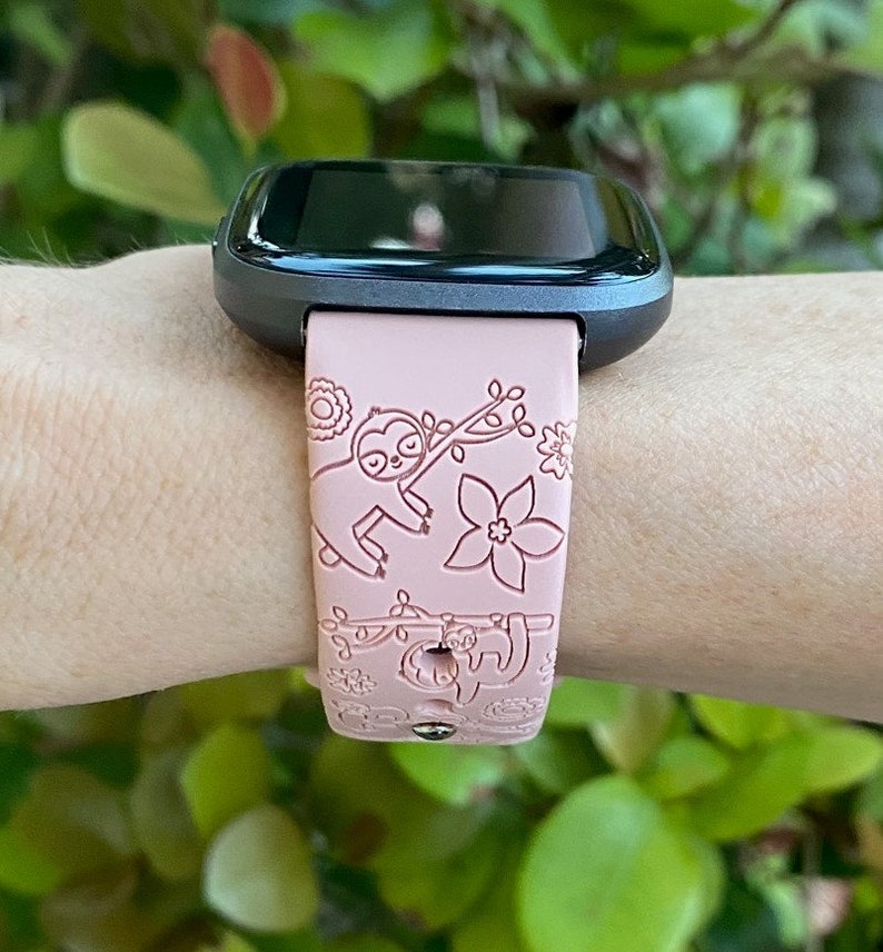 May include: A pink smartwatch band with a sloth design. The band has a black watch face and is worn on a person's wrist.