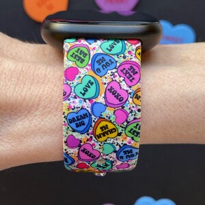 Puede incluir: Un reloj inteligente negro con una banda colorida que presenta un patr&oacute;n de corazones de caramelo con varios mensajes, que incluyen "You B Me", "Text Me", "Dream Big", "Charm Me" y "Love".