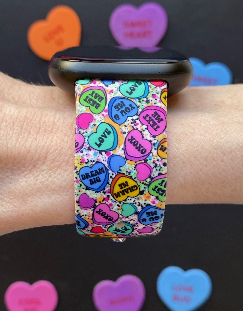 Puede incluir: Un reloj inteligente negro con una banda colorida que presenta un patr&oacute;n de corazones de caramelo con varios mensajes, que incluyen "You B Me", "Text Me", "Dream Big", "Charm Me" y "Love".