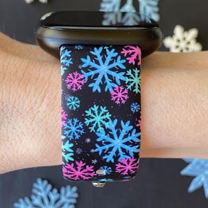 K&ouml;nnte beinhalten: Eine schwarze Smartwatch mit einem schwarzen und wei&szlig;en Silikonarmband mit einem Schneeflockenmuster in Blau, Rosa und T&uuml;rkis.