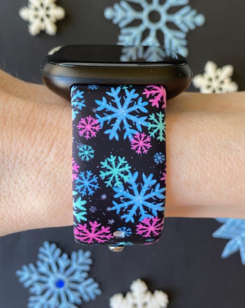 K&ouml;nnte beinhalten: Eine schwarze Smartwatch mit einem schwarzen und wei&szlig;en Silikonarmband mit einem Schneeflockenmuster in Blau, Rosa und T&uuml;rkis.