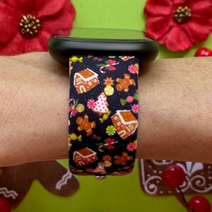 May include: A black smartwatch with a festive band featuring gingerbread houses, gingerbread men, candy canes, and other holiday treats. The band is patterned with a repeating design in shades of brown, red, pink, yellow, and green.