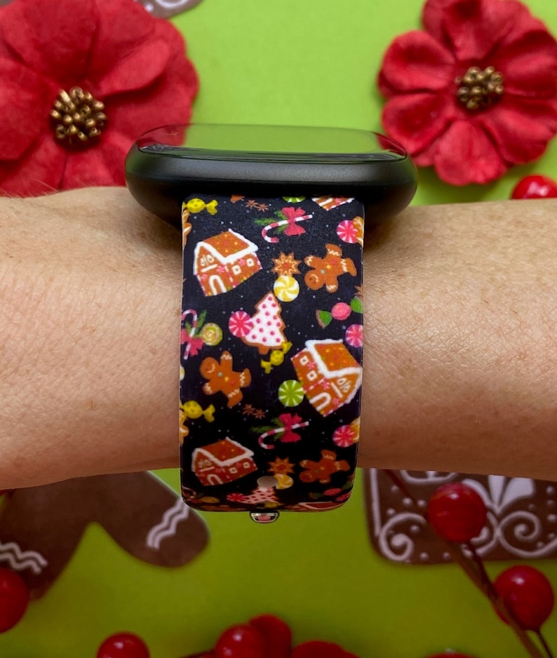 May include: A black smartwatch with a festive band featuring gingerbread houses, gingerbread men, candy canes, and other holiday treats. The band is patterned with a repeating design in shades of brown, red, pink, yellow, and green.