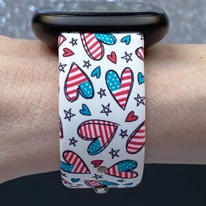 May include: A white watch strap with a patriotic design featuring red and blue hearts, stars, and stripes. The strap is attached to a black smartwatch. The design is reminiscent of the American flag.