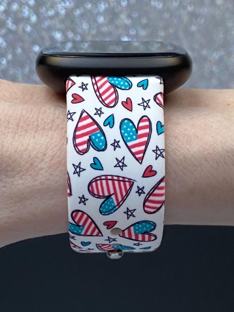 May include: A white watch strap with a patriotic design featuring red and blue hearts, stars, and stripes. The strap is attached to a black smartwatch. The design is reminiscent of the American flag.