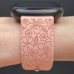 May include: A pink silicone watch band with a floral pattern. The band is designed for a smartwatch with a black face.