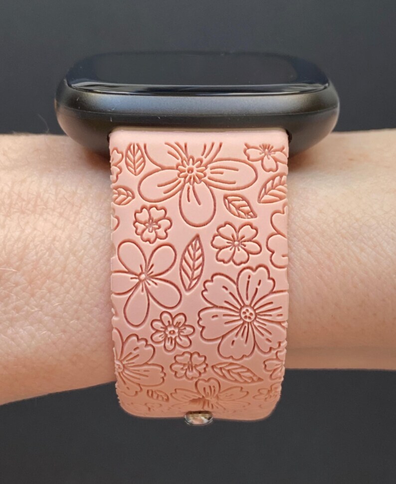 May include: A pink silicone watch band with a floral pattern. The band is designed for a smartwatch with a black face.