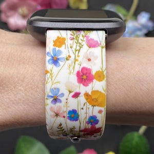 May include: A white watch band with a colorful floral pattern featuring blue, pink, and yellow flowers. The band is attached to a black smartwatch. The design includes a variety of wildflowers.
