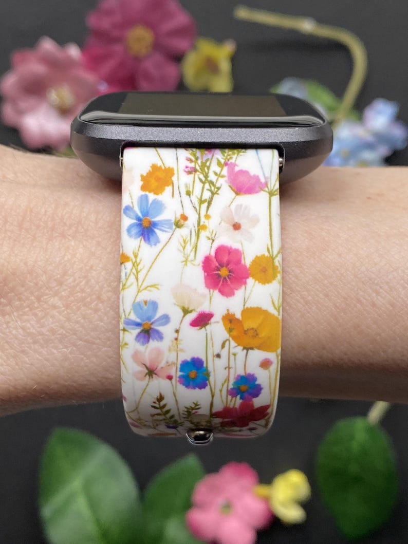 May include: A white watch band with a colorful floral pattern featuring blue, pink, and yellow flowers. The band is attached to a black smartwatch. The design includes a variety of wildflowers.