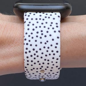 Pu&ograve; includere: Uno smartwatch nero con un cinturino bianco con un motivo a piccoli pois neri distanziati uniformemente. L'orologio &egrave; fissato al polso, mettendo in risalto il design del cinturino e l'estetica moderna dell'orologio.