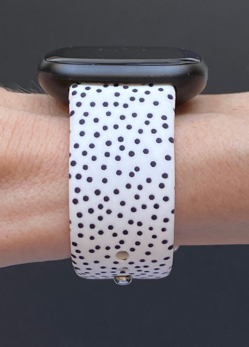 Pu&ograve; includere: Uno smartwatch nero con un cinturino bianco con un motivo a piccoli pois neri distanziati uniformemente. L'orologio &egrave; fissato al polso, mettendo in risalto il design del cinturino e l'estetica moderna dell'orologio.