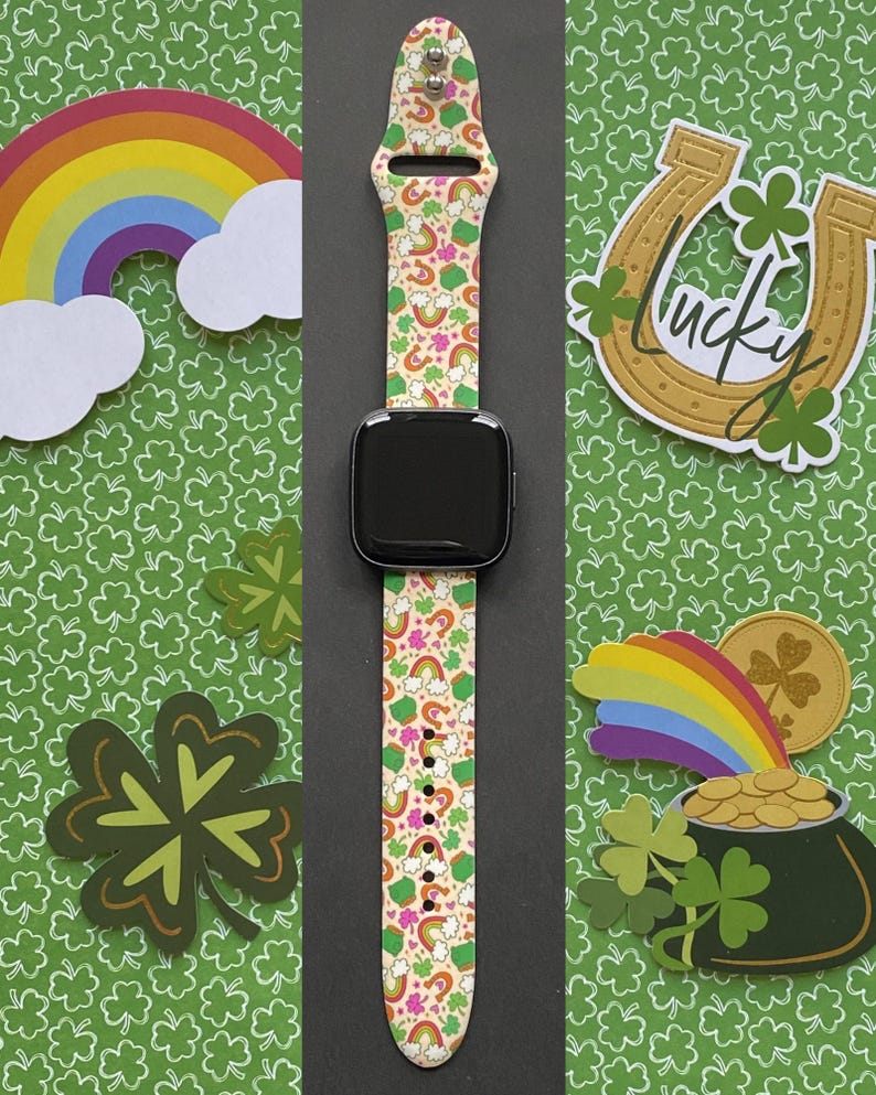 May include: A smartwatch with a cream-colored band featuring rainbows, shamrocks, and unicorns. The watch is set against a green background with shamrock cutouts and other St. Patrick's Day themed decorations.