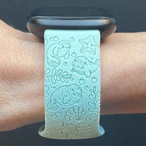 May include: A light blue watch band with a sea life design. The band features engraved illustrations of manatees, starfish, bubbles, seaweed, and seashells. The watch band is attached to a black watch.