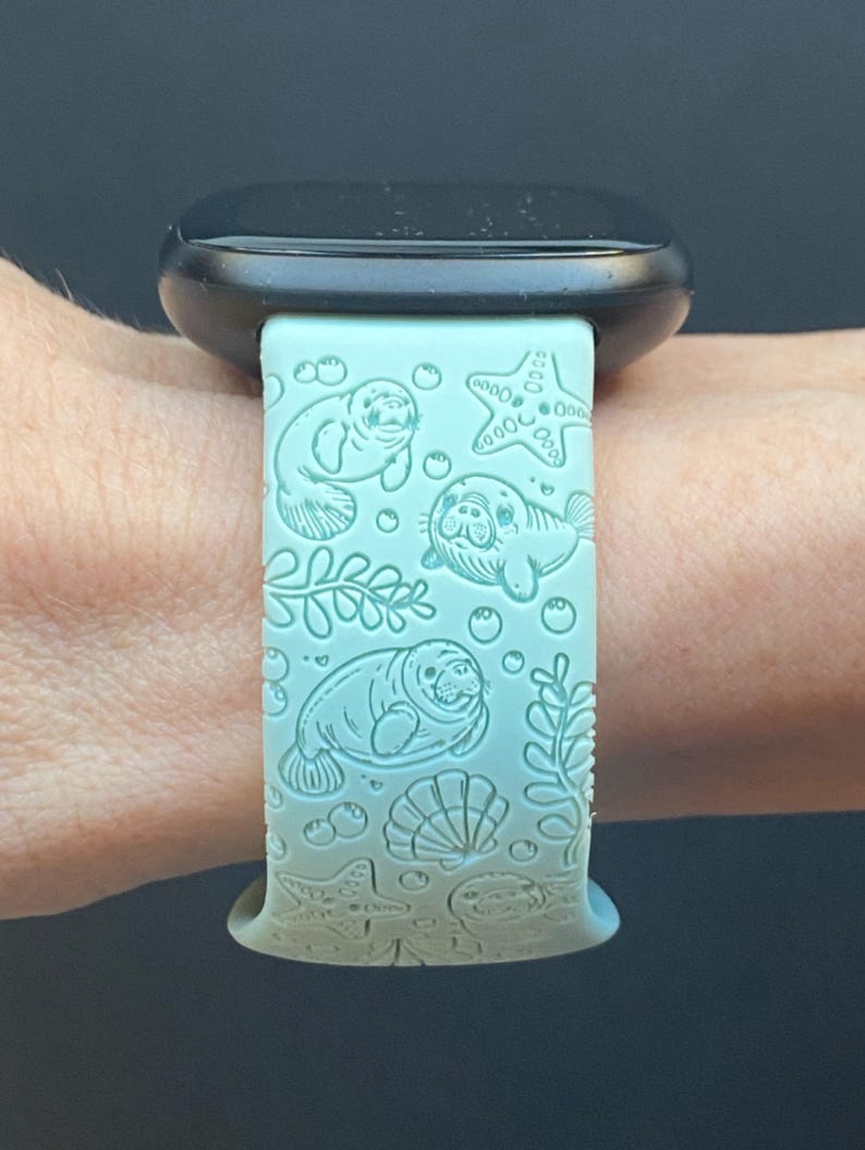 May include: A light blue watch band with a sea life design. The band features engraved illustrations of manatees, starfish, bubbles, seaweed, and seashells. The watch band is attached to a black watch.