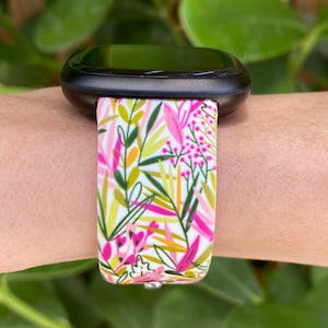 May include: A white smartwatch band with a floral pattern featuring pink, green, and yellow leaves. The band is made of silicone and is compatible with most smartwatches.