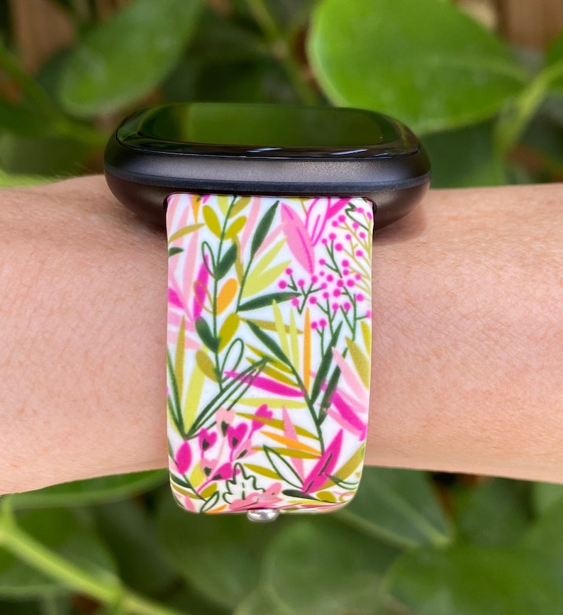 May include: A white smartwatch band with a floral pattern featuring pink, green, and yellow leaves. The band is made of silicone and is compatible with most smartwatches.