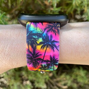 May include: A black smartwatch with a vibrant watch band featuring a tropical palm tree design. The band showcases a colorful gradient of pink, yellow, and blue hues, with black palm tree silhouettes. The watch is attached to a wrist.
