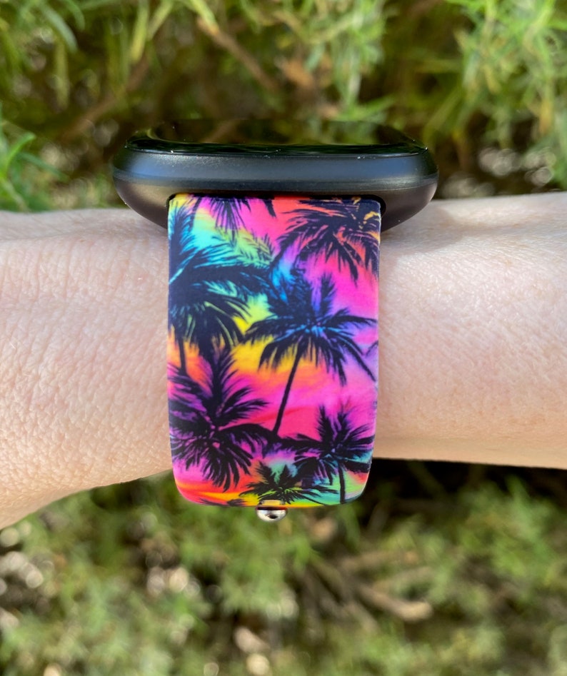 May include: A black smartwatch with a vibrant watch band featuring a tropical palm tree design. The band showcases a colorful gradient of pink, yellow, and blue hues, with black palm tree silhouettes. The watch is attached to a wrist.
