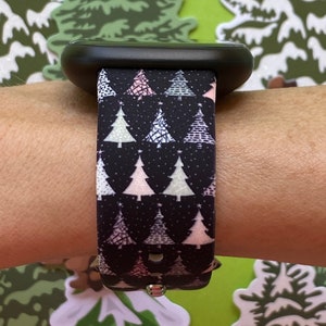 May include: A black smartwatch band with a Christmas tree pattern. The band has a white background with pink, green, and white Christmas trees. The trees are outlined in black and have a white star on top.