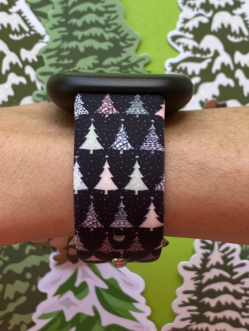 May include: A black smartwatch band with a Christmas tree pattern. The band has a white background with pink, green, and white Christmas trees. The trees are outlined in black and have a white star on top.