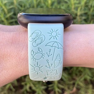 May include: A light green watch band with a beach-themed design. The band features a variety of icons, including flip-flops, a sun, an umbrella, a crab, an anchor, a seashell, and a sandcastle.