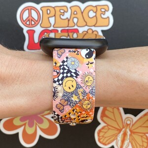 Op de afbeelding: Een roze en oranje Apple Watch band met een kleurrijk patroon van vlinders, zonnen, sterren en smiley-gezichten. De band is gemaakt van siliconen en heeft een zwarte gesp.