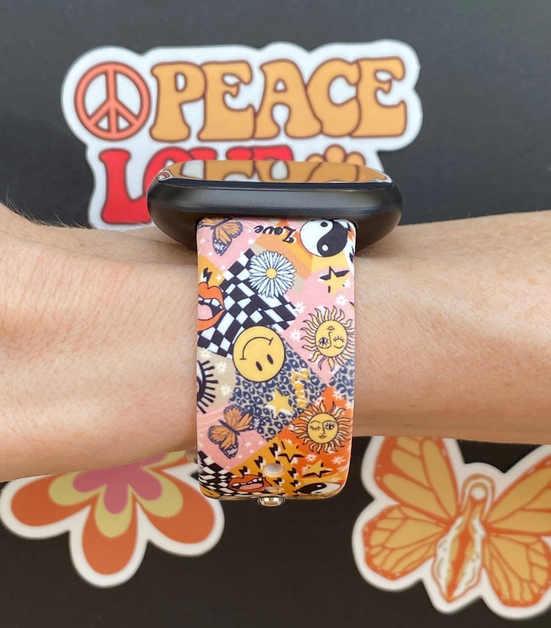 Op de afbeelding: Een roze en oranje Apple Watch band met een kleurrijk patroon van vlinders, zonnen, sterren en smiley-gezichten. De band is gemaakt van siliconen en heeft een zwarte gesp.