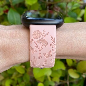 May include: A pink silicone watch band with a floral and butterfly design. The band is compatible with smartwatches.