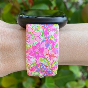 K&ouml;nnte beinhalten: Eine schwarze Smartwatch mit einem pink- und gr&uuml;n gemusterten Armband. Das Armband hat einen wei&szlig;en Hintergrund mit pinken, gr&uuml;nen und violetten Blumen.