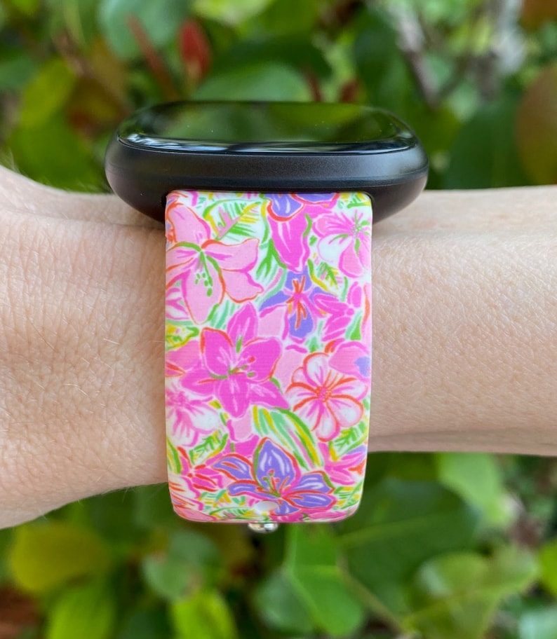 K&ouml;nnte beinhalten: Eine schwarze Smartwatch mit einem pink- und gr&uuml;n gemusterten Armband. Das Armband hat einen wei&szlig;en Hintergrund mit pinken, gr&uuml;nen und violetten Blumen.