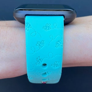May include: A turquoise blue fitness tracker band with a bee pattern. The band is made of silicone and has a small red dot on the bottom.