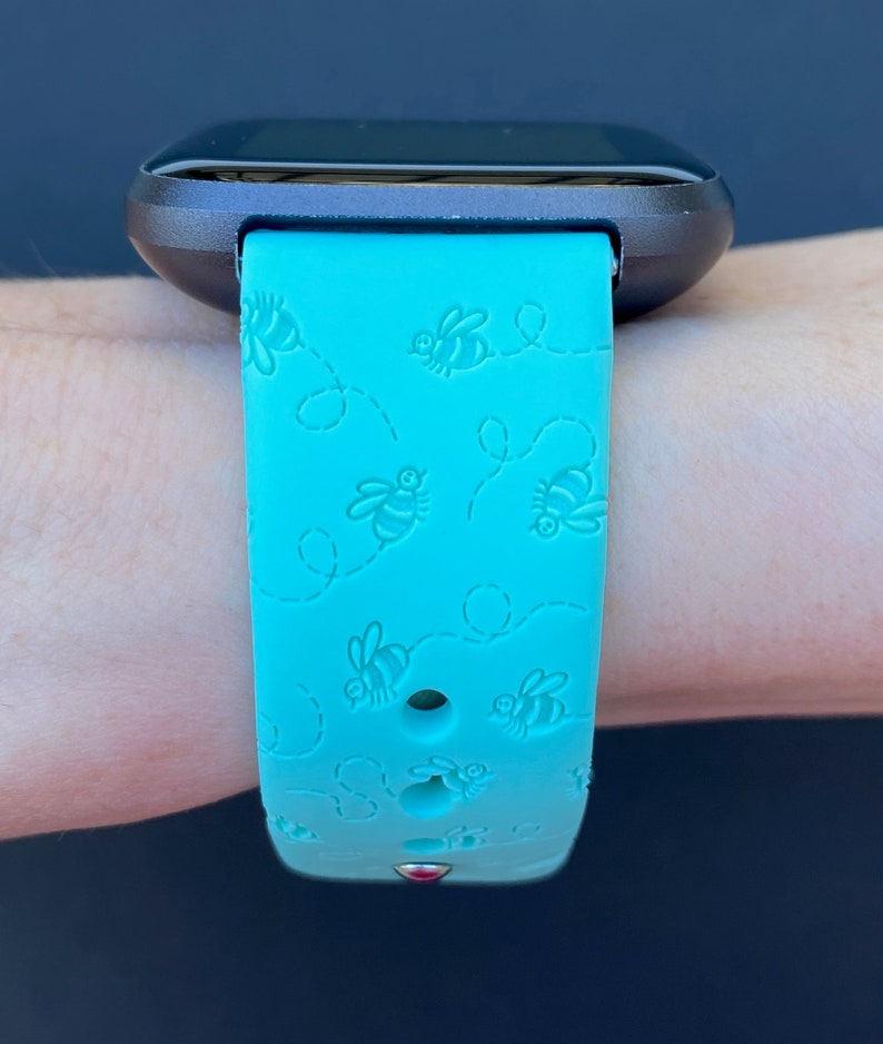May include: A turquoise blue fitness tracker band with a bee pattern. The band is made of silicone and has a small red dot on the bottom.