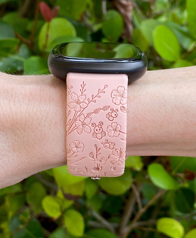 May include: A pink silicone watch band with a floral and bee design. The band is shown on a person's wrist.