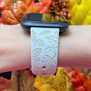 May include: A smartwatch with a cream-colored band featuring an embossed autumn leaf and acorn design. The watch has a dark gray bezel and a black screen. The band is secured on a wrist.