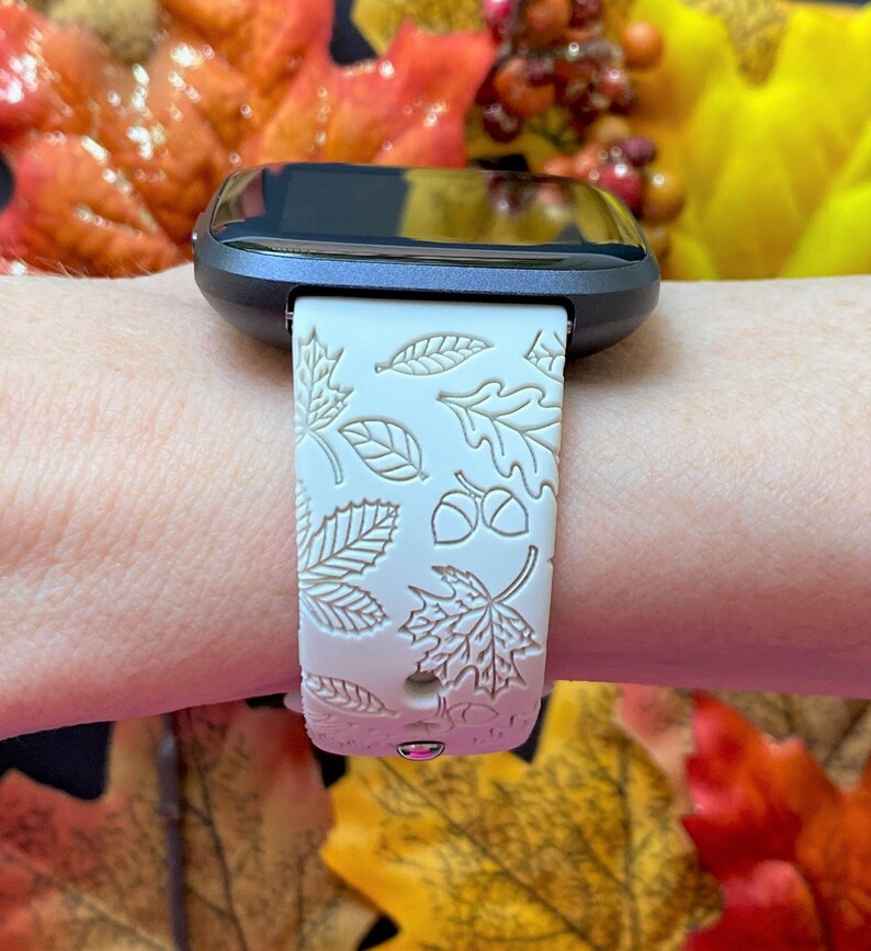 May include: A smartwatch with a cream-colored band featuring an embossed autumn leaf and acorn design. The watch has a dark gray bezel and a black screen. The band is secured on a wrist.