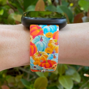 K&ouml;nnte beinhalten: Eine schwarze Smartwatch mit einem bunten Armband mit K&uuml;rbis- und Blumenmuster. Das Armband ist aus Silikon und hat eine leuchtend orange, blaue und gelbe Farbgebung.