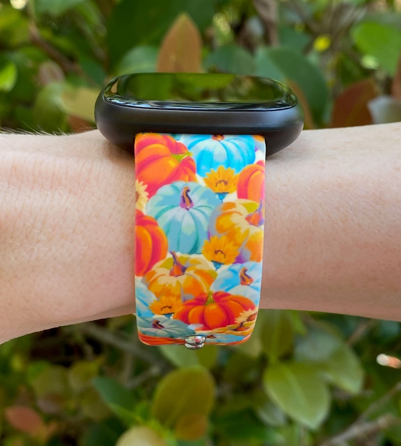 K&ouml;nnte beinhalten: Eine schwarze Smartwatch mit einem bunten Armband mit K&uuml;rbis- und Blumenmuster. Das Armband ist aus Silikon und hat eine leuchtend orange, blaue und gelbe Farbgebung.