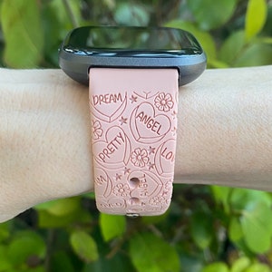 May include: A pink smartwatch strap featuring embossed hearts, flowers, and the words "DREAM", "PRETTY", "ANGEL", and "LOVE". The strap is attached to a black smartwatch face.
