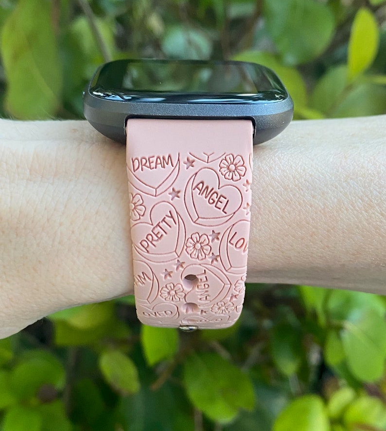 May include: A pink smartwatch strap featuring embossed hearts, flowers, and the words "DREAM", "PRETTY", "ANGEL", and "LOVE". The strap is attached to a black smartwatch face.
