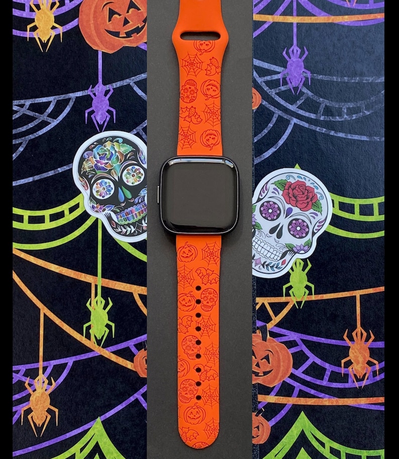 May include: Orange smartwatch band with a Halloween-themed design featuring skulls, pumpkins, and spiderwebs. The band is displayed against a black background with Halloween decorations. The watch face is black.