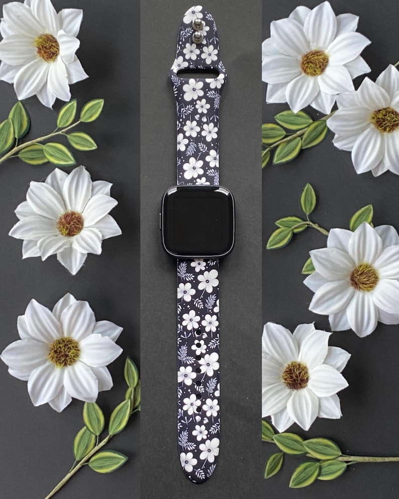 May include: A black smartwatch with a floral patterned band featuring white flowers and leaves. The watch is displayed on a dark background, surrounded by white flowers and green leaves.