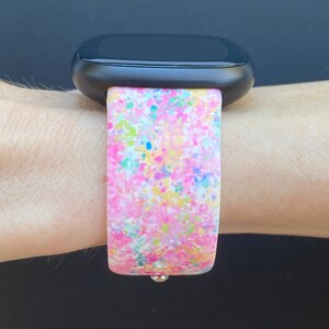May include: A pink, white, blue, and yellow splatter pattern watch band for a smartwatch.