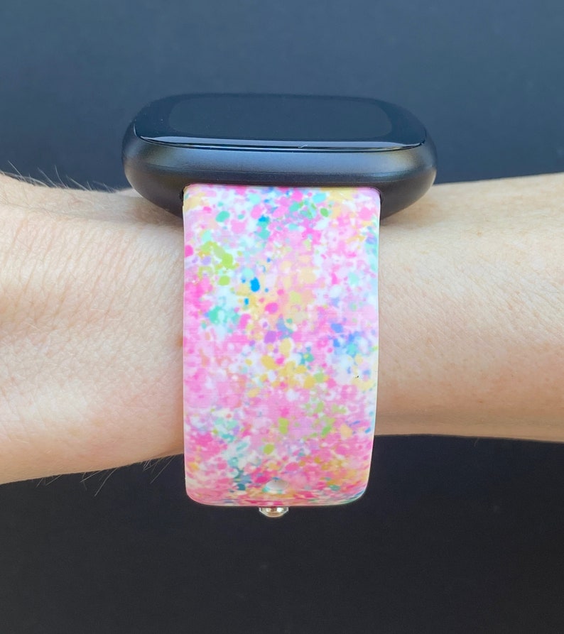 May include: A pink, white, blue, and yellow splatter pattern watch band for a smartwatch.