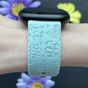 May include: A light blue watch band with the words "Grow in Faith, Faith over Fear" and floral and butterfly designs. The watch band is attached to a black watch face. The image includes purple and yellow flowers.