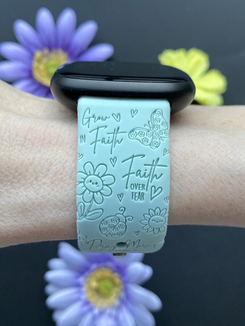 May include: A light blue watch band with the words "Grow in Faith, Faith over Fear" and floral and butterfly designs. The watch band is attached to a black watch face. The image includes purple and yellow flowers.