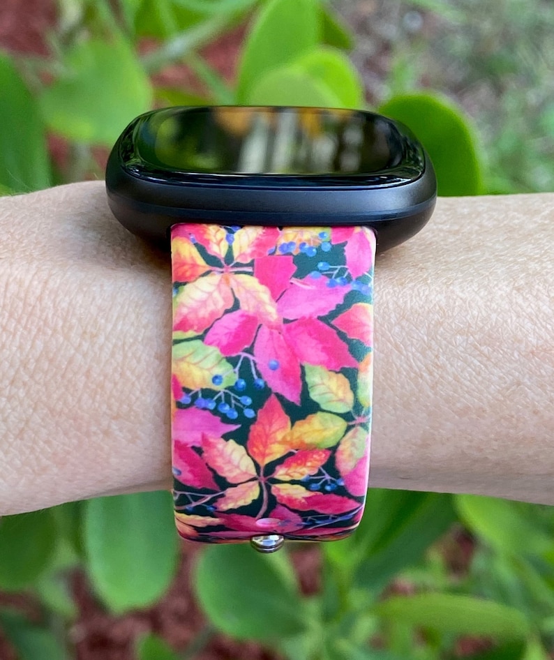 May include: A black smartwatch with a colorful floral band. The band features a pink, yellow, and green floral pattern with dark green leaves and small blue berries.