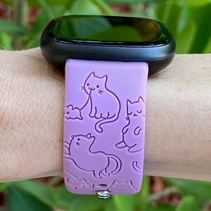 May include: A purple silicone watch band with a cat design. The band has a repeating pattern of cats in different poses, including a cat licking its paw, a cat sitting, and a cat sleeping. The band is compatible with smartwatches.