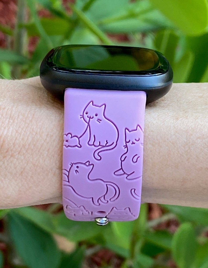 May include: A purple silicone watch band with a cat design. The band has a repeating pattern of cats in different poses, including a cat licking its paw, a cat sitting, and a cat sleeping. The band is compatible with smartwatches.