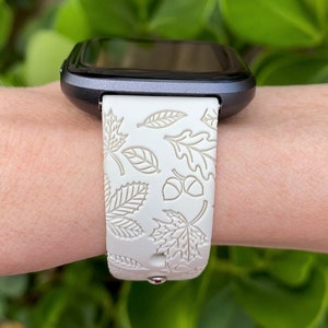 May include: A smartwatch with a white band featuring a leaf and acorn pattern. The band is made of a flexible material and is attached to a dark gray watch face. The design evokes an autumn theme.